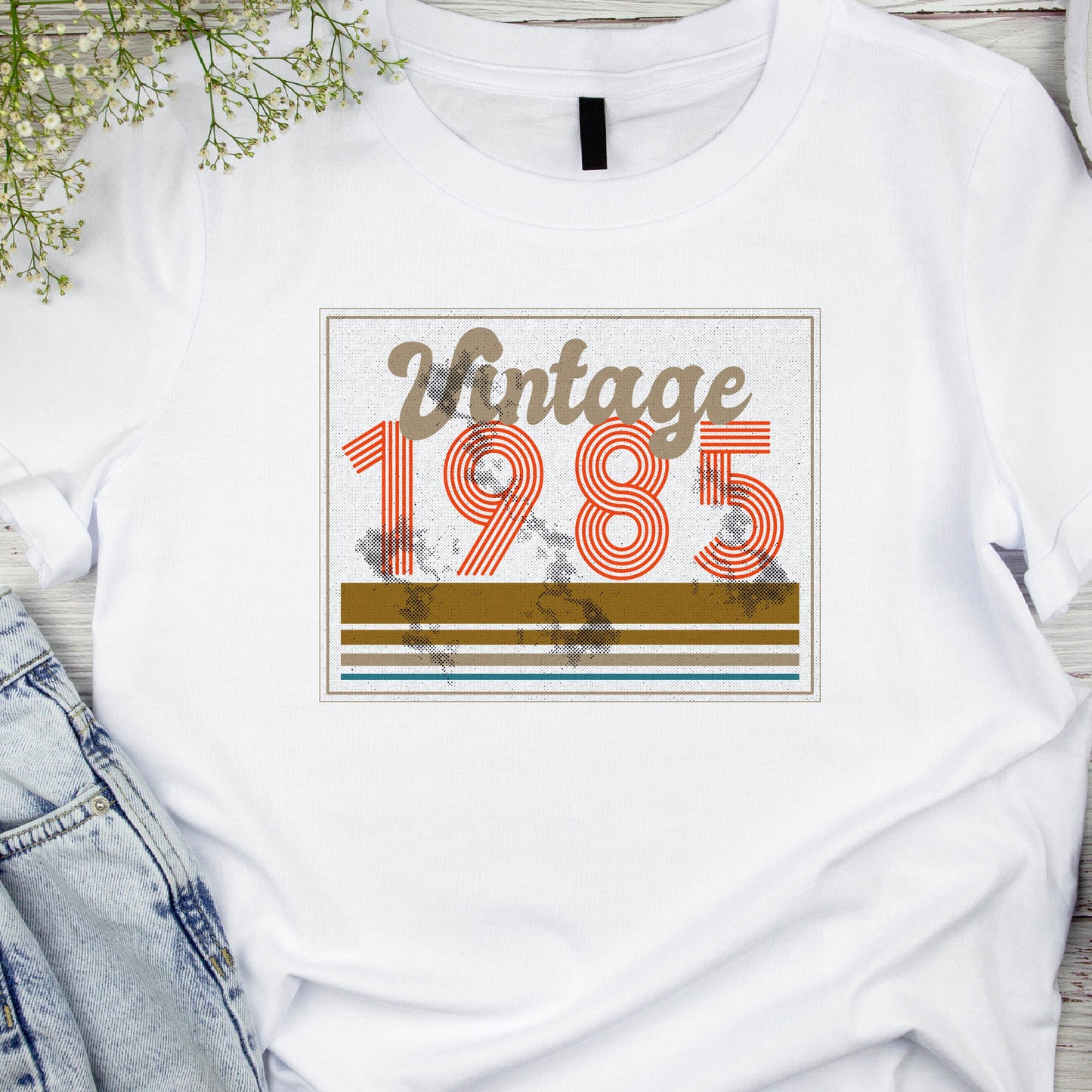 White crew neck t-shirt with the text 'Vintage 1985' printed on the front, displayed on a wooden surface with denim jeans and a pair of shoes in the background.
