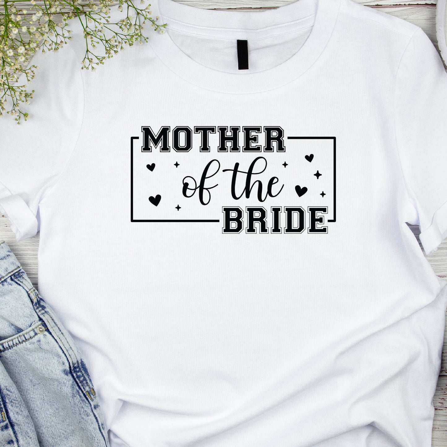 A white t-shirt with the text 'MOTHER OF THE BRIDE' printed on the front, styled with a pair of blue jeans and casual footwear in a casual setting, likely with decorative elements in the background.