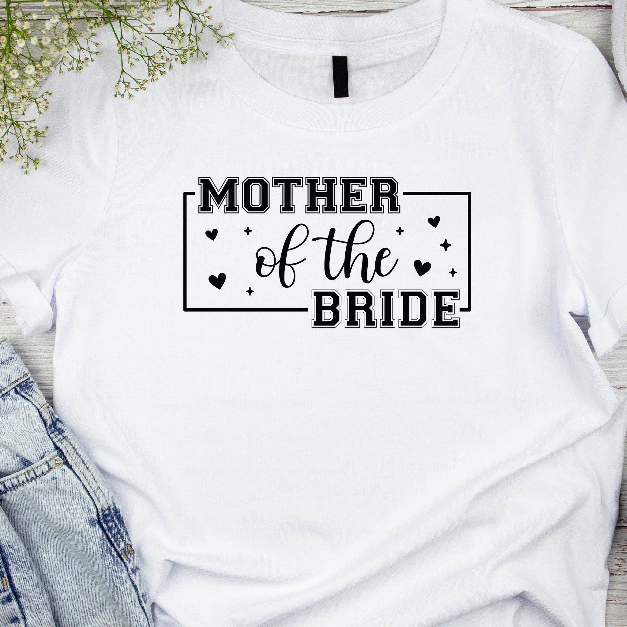 A white t-shirt with the text 'MOTHER OF THE BRIDE' printed on the front, styled with a pair of blue jeans and casual footwear in a casual setting, likely with decorative elements in the background.