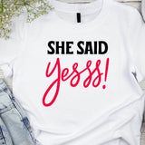 White t-shirt with the text 'SHE SAID YEEE!' in red, bold, uppercase letters, positioned on a table with denim jeans and a black object, possibly a shoe, in the background.
