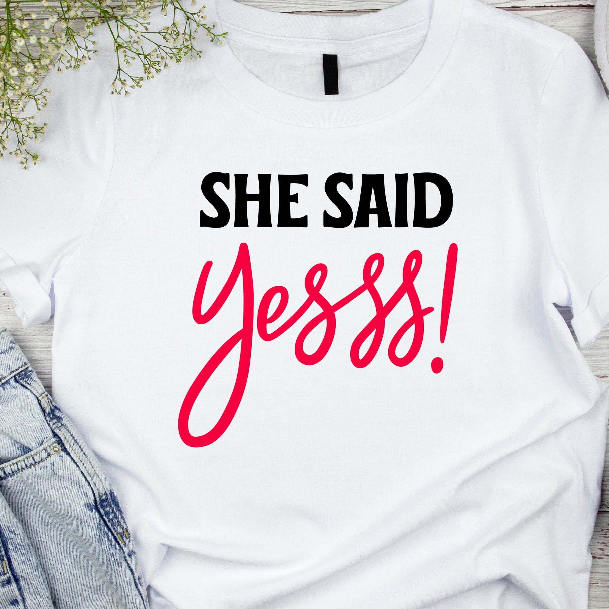 White t-shirt with the text 'SHE SAID YEEE!' in red, bold, uppercase letters, positioned on a table with denim jeans and a black object, possibly a shoe, in the background.