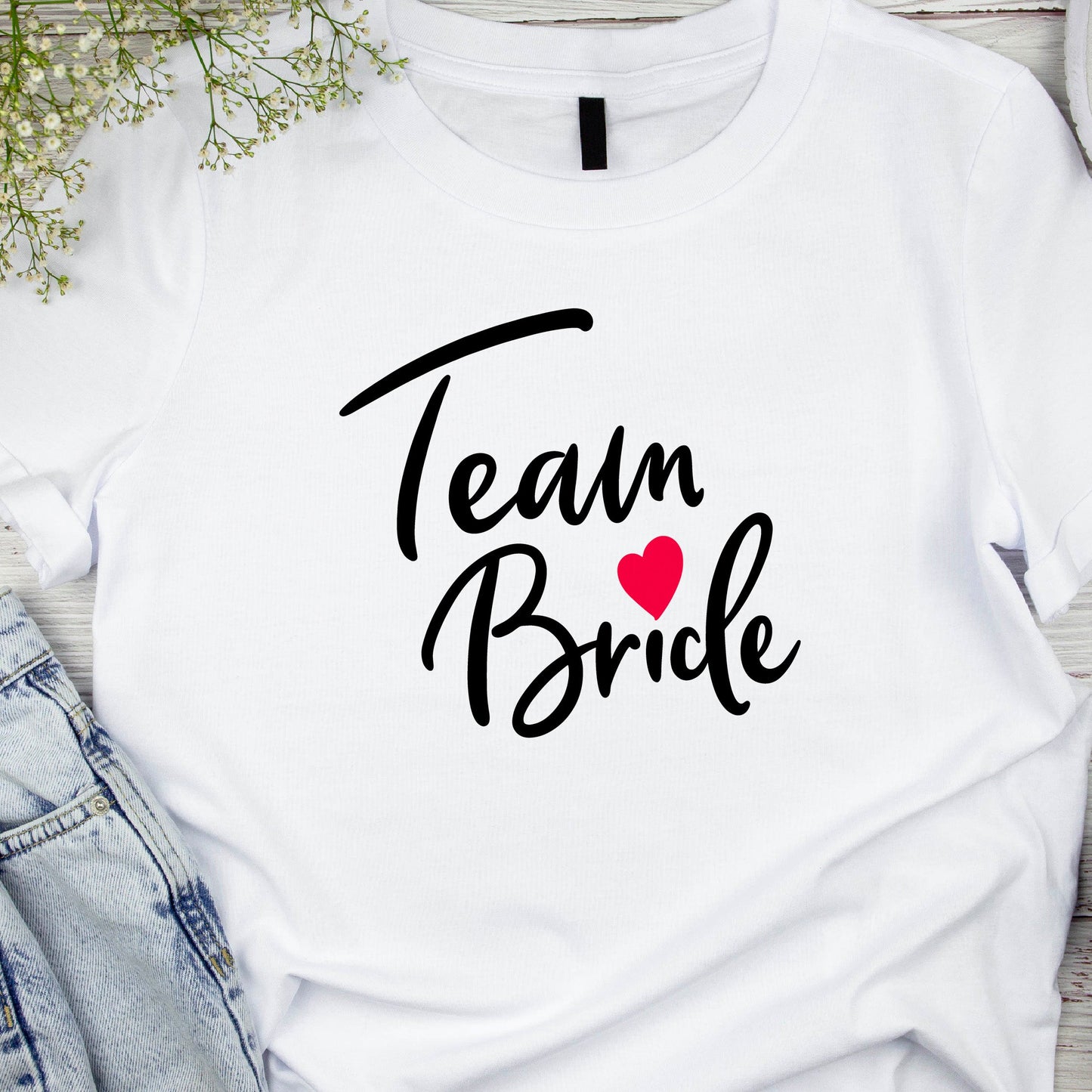 A white crew neck t-shirt with the text 'Team Bride' printed in black, accompanied by a red heart. The shirt is displayed flat with a pair of blue jeans and a pair of black heels in the background.