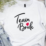 A white crew neck t-shirt with the text 'Team Bride' printed in black, accompanied by a red heart. The shirt is displayed flat with a pair of blue jeans and a pair of black heels in the background.