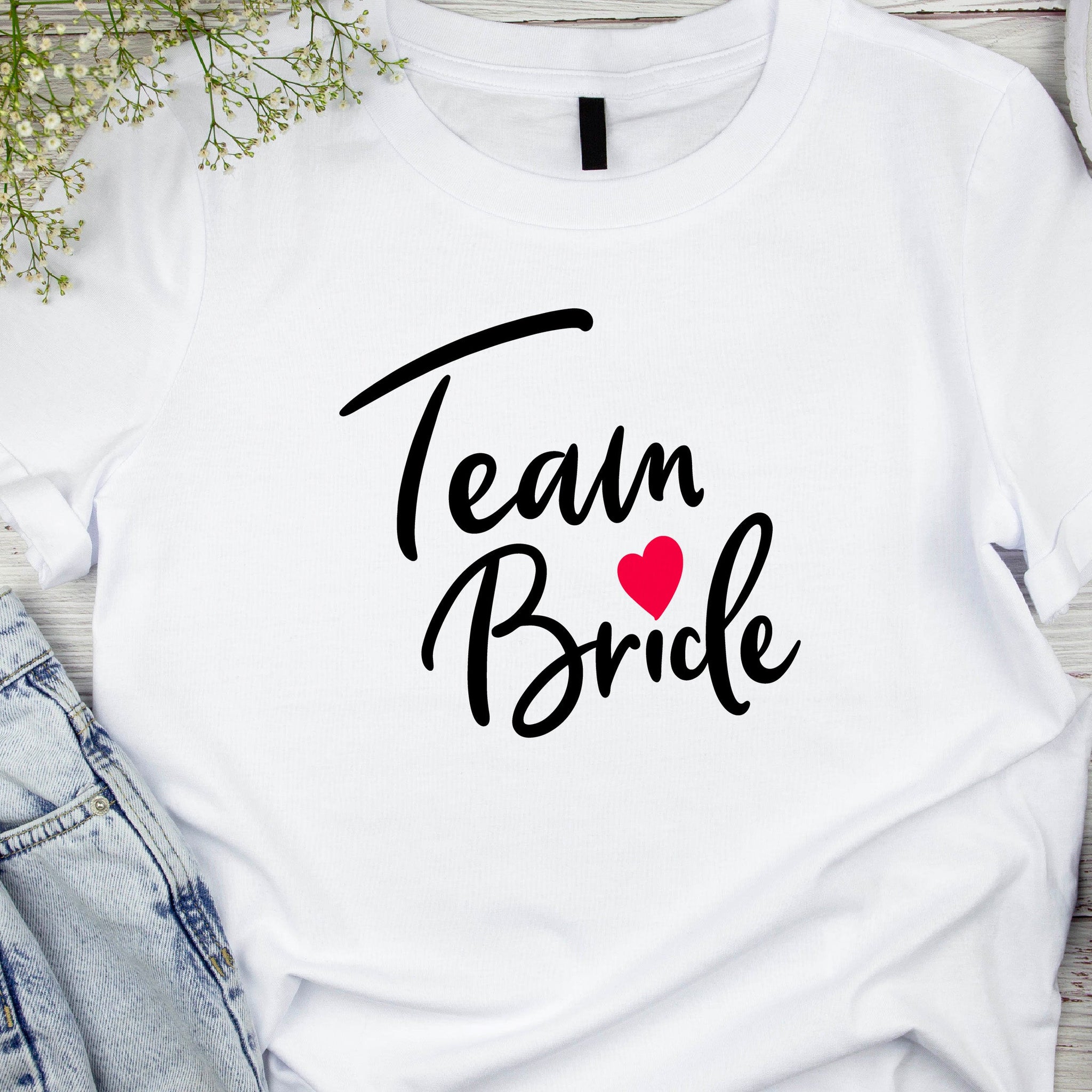 A white crew neck t-shirt with the text 'Team Bride' printed in black, accompanied by a red heart. The shirt is displayed flat with a pair of blue jeans and a pair of black heels in the background.
