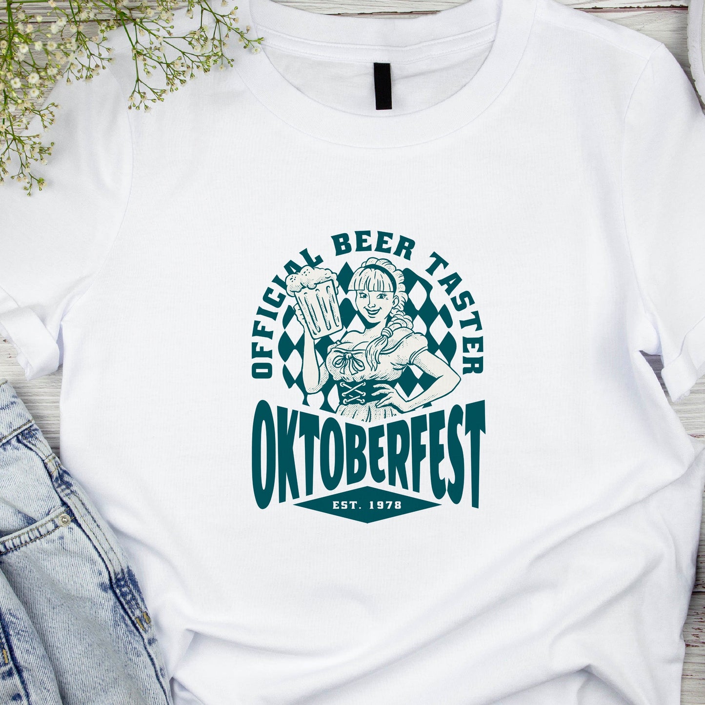White t-shirt with Oktoberfest design featuring a person holding a beer mug, placed on a wooden surface with denim jeans and headphones.