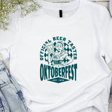 White t-shirt with Oktoberfest design featuring a person holding a beer mug, placed on a wooden surface with denim jeans and headphones.