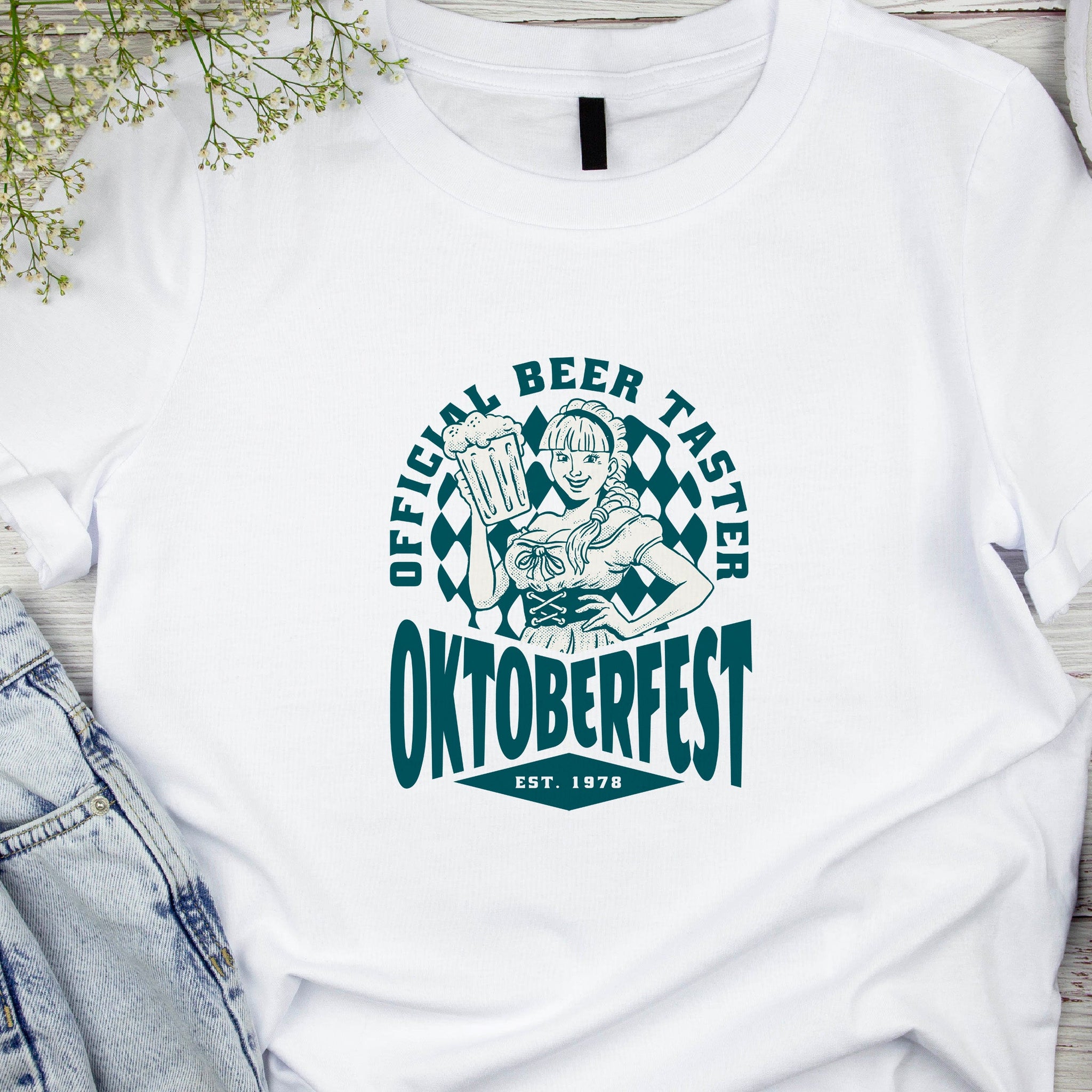 White t-shirt with Oktoberfest design featuring a person holding a beer mug, placed on a wooden surface with denim jeans and headphones.